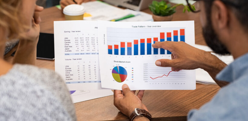 Mature latin businessman showing stock market bar graph to businesswoman. Financial advising team discussing trading patterns of year. Rear view of casual businesspeople holding charts studying year trend together.